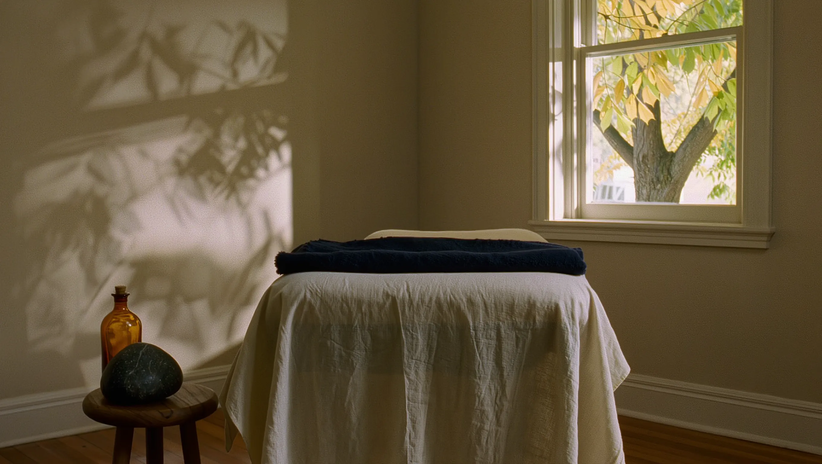 A massage table made up with cream linen and a folded navy blanket, lit by afternoon sun through a window with a tree outside.
