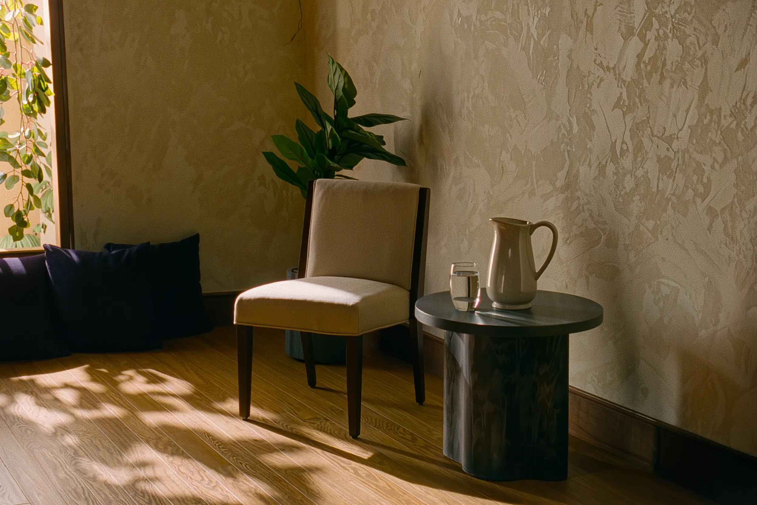 A quiet corner with a wooden chair, a pitcher of water and a potted plant, warm light falling across cream walls.